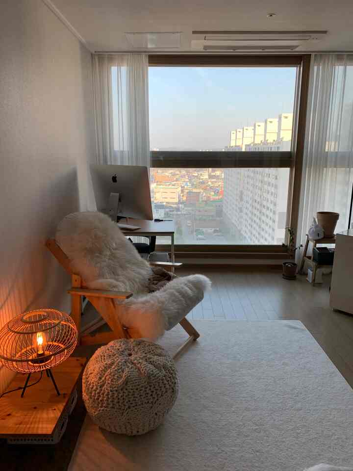 Natural tone studio apartment featuring a desk by large window with white chiffon curtains, wooden armchair with sheepskin, and knit pouf creating a cozy atmosphere