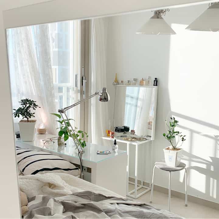 Bright white-toned bedroom with curtained windows, vanity mirror, desk lamp, and plants creating a cozy atmosphere