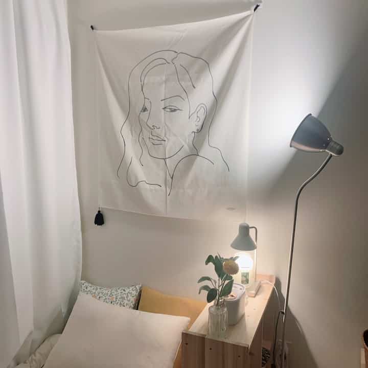 Bright white-toned bedroom featuring a bed with cushions next to a wooden shelf and floor lamp creating a cozy atmosphere