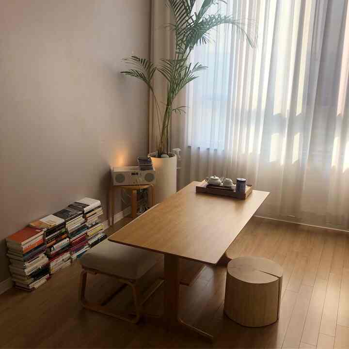 Natural wood-tone dining room featuring a wooden table, stools, sheer curtains, plant, and stacked books creating warm atmosphere