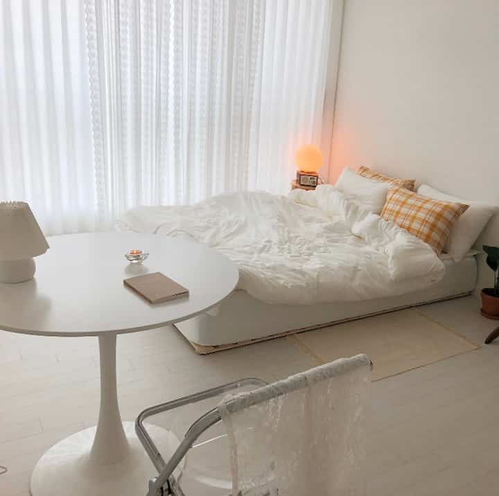 A modern bedroom in white and beige tones featuring a round table, cushions, and light curtains creating a cozy atmosphere in a small room