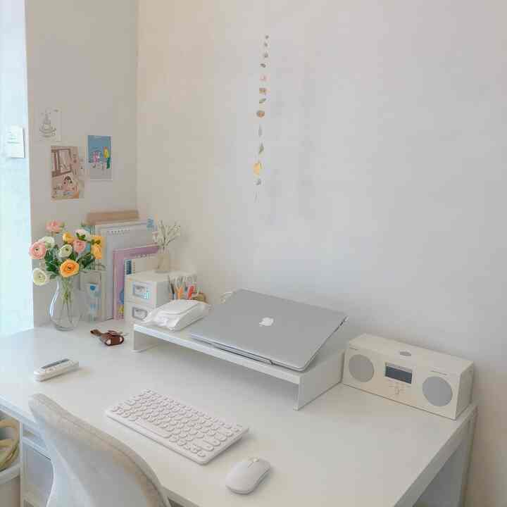 White-toned home office space featuring a tidy desk with laptop, Bluetooth speaker, and vase with flowers creating a cozy workspace