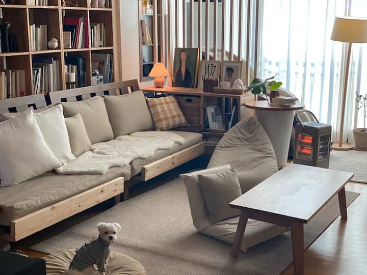 A living room with wood tones and beige accents featuring a wooden sofa, bookshelf, rug, bean bag, and a small pet dog in a cozy setting