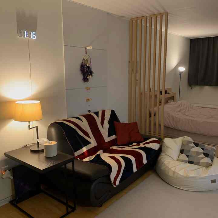 Natural wood tones and black sofa in a studio apartment with a living room and bedroom separated by wooden partition, featuring soft lighting and blackout curtains for a cozy atmosphere