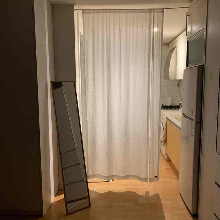 A cozy studio apartment entrance and kitchen separated by white curtain featuring wood tone flooring and cabinetry