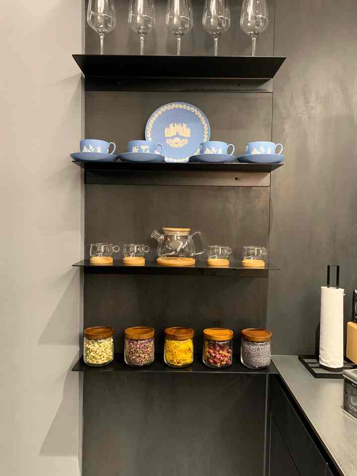 ブラックのメタル棚にラベンダーカラーのティーカップセットと透明なガラス製のティーポット、さまざまなハーブの瓶が配置されたモダンなキッチン空間