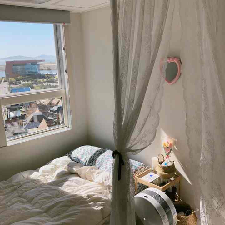 A white-toned bedroom with a single bed and lace curtains by the window, flooded with natural light creating a cozy atmosphere