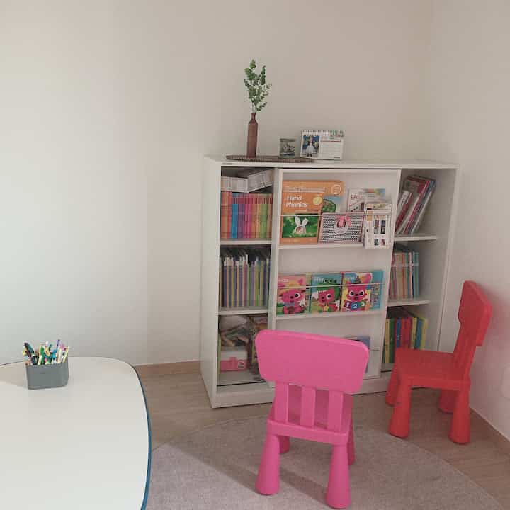 Simple white-toned kids' room featuring pink and red child chairs and a bookshelf filled with colorful books