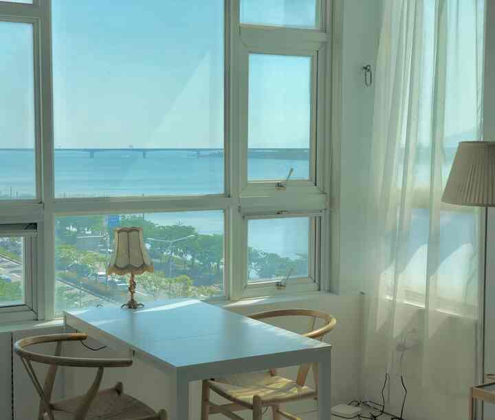 Bright white and wood tone dining room featuring large windows showing a sea view, with a soft table lamp creating a natural atmosphere