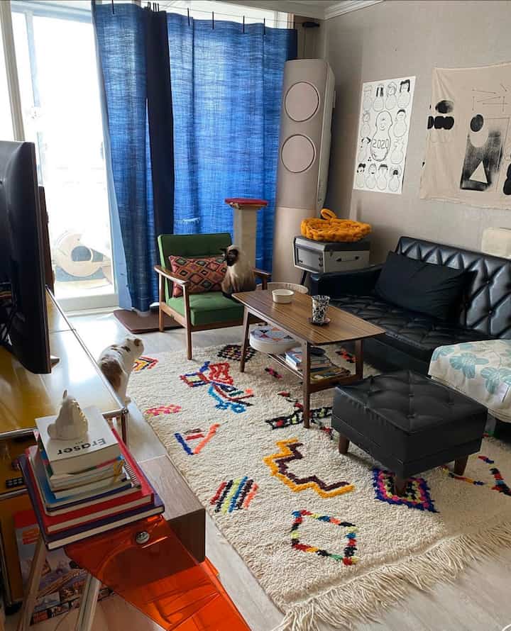 青いカーテンとカラフルな柄のラグがあるリビング、黒いソファと緑のアームチェア、2匹の猫がいる落ち着いた雰囲気の空間