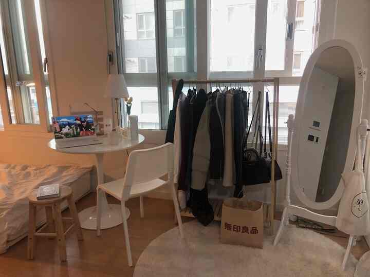 Natural color studio apartment bedroom featuring bed, round table with laptop, clothing rack, and full-length mirror in a cozy space