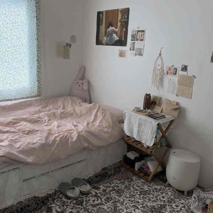 パステルピンクの寝具とヴィンテージのペルシャ絨毯が印象的なワンルームの寝室、自然光と温かみのあるインテリアが調和した空間