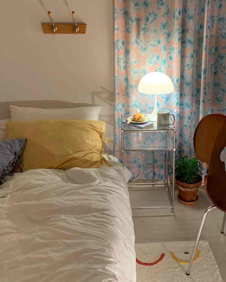 A cozy bedroom in soft white and yellow tones featuring a metal nightstand and a table lamp with floral patterned curtains