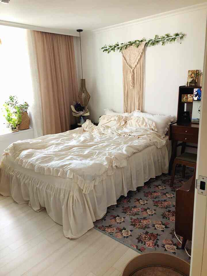 アイボリーとライトブラウンのトーンの寝室、フリル付き寝具と化粧台、花柄ラグ、ペット用スクラッチャーとベッドが配置された雰囲気のあるインテリア