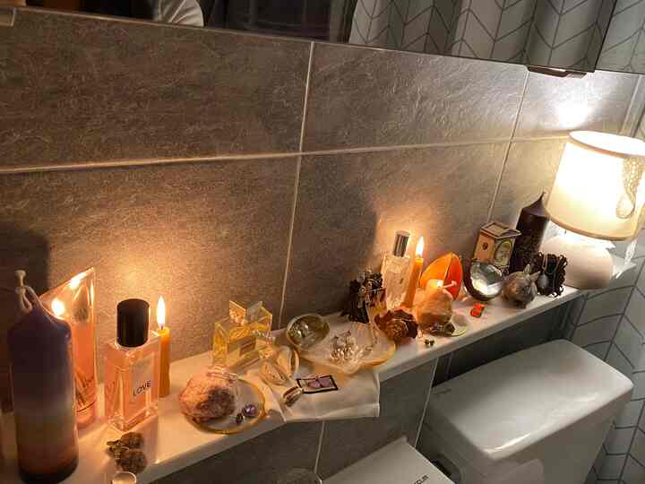 Bathroom with gray tiled walls and white toilet featuring vintage items, scented candles, candles, and a table lamp on the shelf creating a warm atmosphere