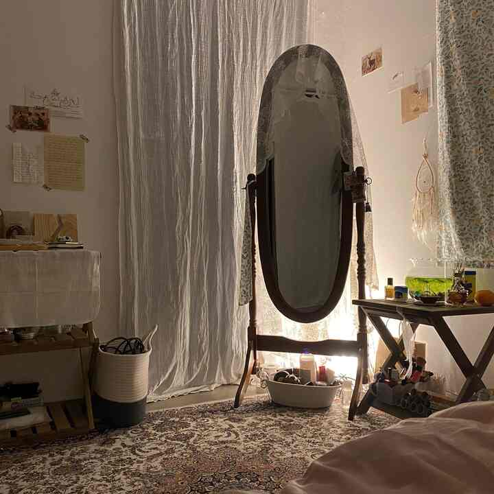 柔らかなナチュラルトーンのワンルーム寝室にヴィンテージのスタンドミラー、ペルシャ絨毯、カーテン、木製ナイトテーブルが配置された雰囲気のあるインテリア