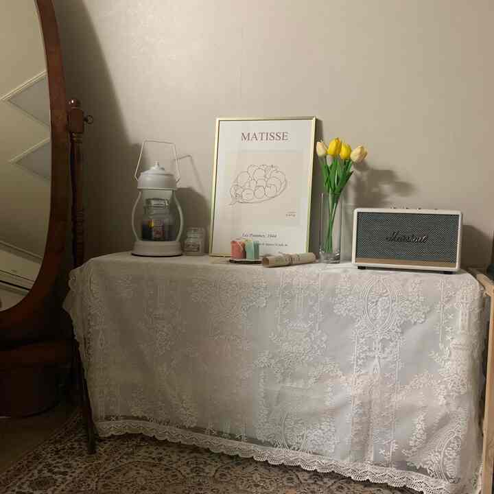 White lace tablecloth-covered bedroom space featuring a central Matisse picture frame and yellow tulip vase, with a wooden mirror positioned left, evoking a cozy atmosphere