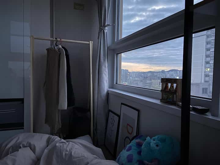 柔らかなグレーとホワイトのトーンの寝室、窓辺に手描きの額縁と洋服掛けがある落ち着いた雰囲気の空間