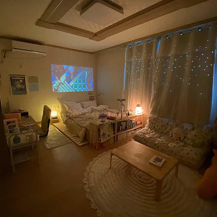 Warm natural-toned studio living room featuring wooden furniture, bed, and projector screen with cozy atmosphere