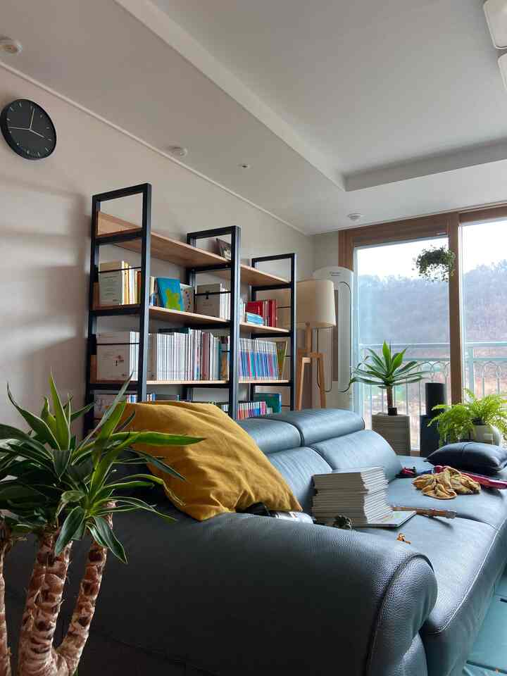 ナチュラルカラーのリビング、木製本棚とダークグレーのソファ、マスタードクッション、多数の植物が配置された雰囲気のあるインテリア