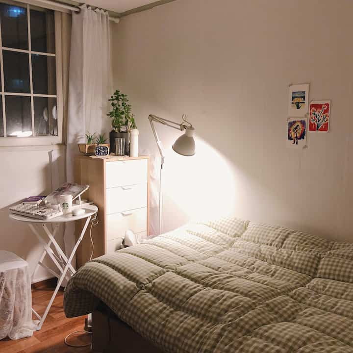 Small cozy bedroom in white and light brown tones featuring checkered bedding, dresser, lamp, and plants with a calming atmosphere