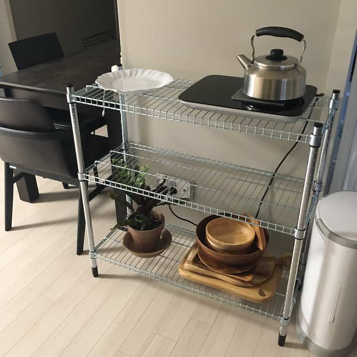 White and natural toned kitchen space with metal shelving holding stainless steel kettle, adjacent dining chairs creating clean look