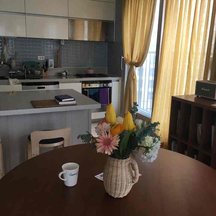 黄色いカーテンが掛かったキッチンとダイニングルーム、丸テーブルの上にラタンの花瓶とコーヒーカップが置かれた雰囲気のある空間