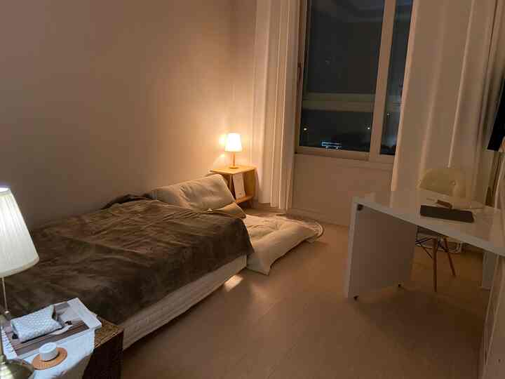 温かい照明とベージュ色調のワンルーム寝室、ベッドとデスクが配置された落ち着いた空間