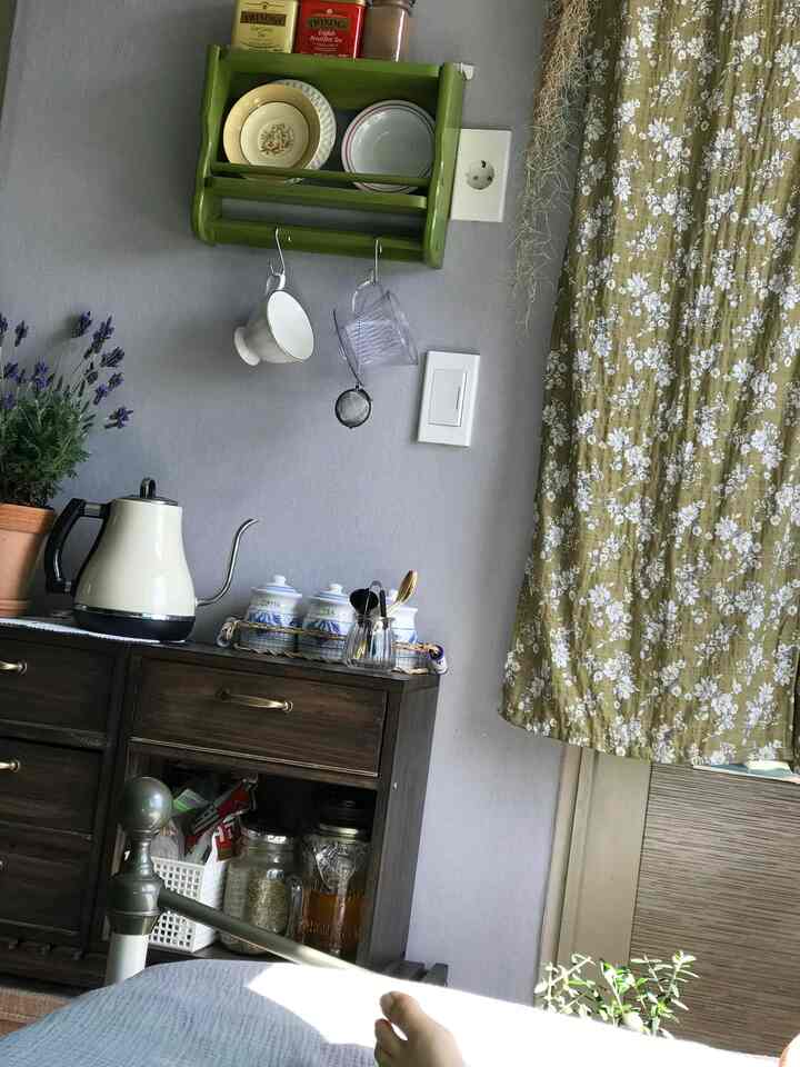 A cozy vintage-style bedroom with light gray walls and olive floral curtains, featuring a dark brown cabinet with coffee drip kettle and tea accessories