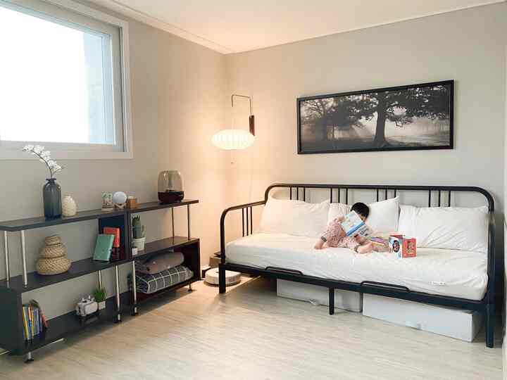 Natural-tone study and bedroom space featuring a black metal bed with white mattress on the right side, a baby reading books on the bed