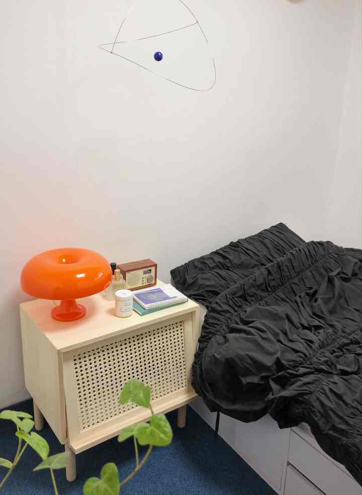A bedroom with white walls and black bedding featuring an orange table lamp on a natural wood nightstand, creating a cozy atmosphere