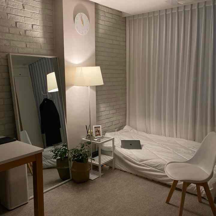 White-toned studio bedroom featuring a full-length mirror, floor lamp, and plant, creating a cozy and neat interior