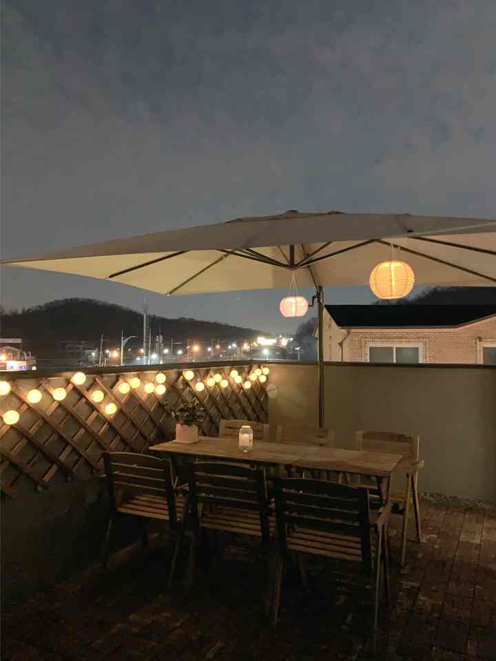 ベージュのトーンのバルコニー空間、木製のアウトドアテーブルと椅子、ストリングライトとペンダントライトが温かみのある雰囲気を演出するインテリア