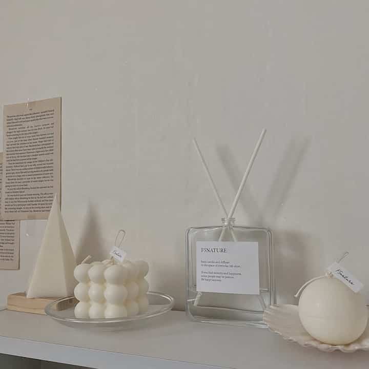 Minimal interior decor featuring various shaped white candles and diffuser arranged on a white shelf with a white wall background