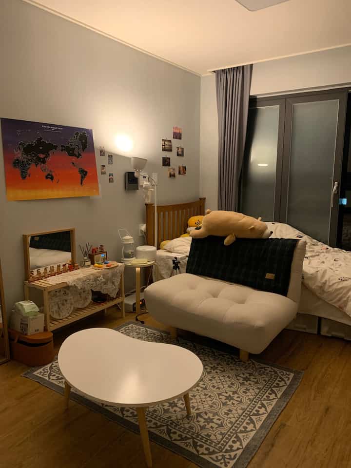 Natural toned small bedroom and living area featuring wooden bed, cream sofa, and white coffee table with cozy atmosphere