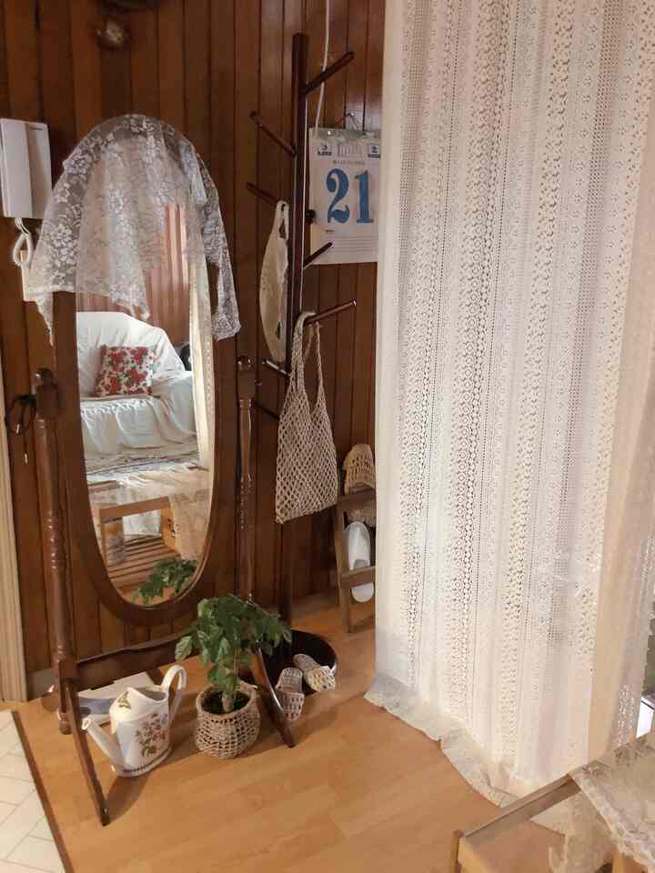 アイボリーのレースカーテンと木目調の壁、全身鏡とコートハンガーが配置されたナチュラルな玄関の空間