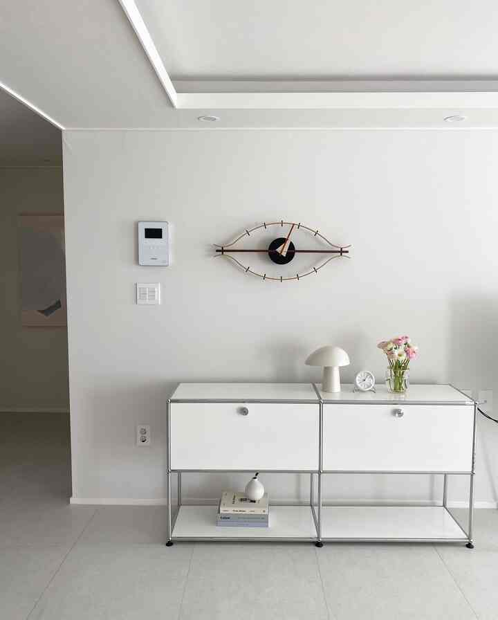明るいホワイトトーンのリビング、中央に白いサイドボードとユニークな目の形の時計、右側には花瓶とテーブルランプが配置された清潔でモダンなインテリア
