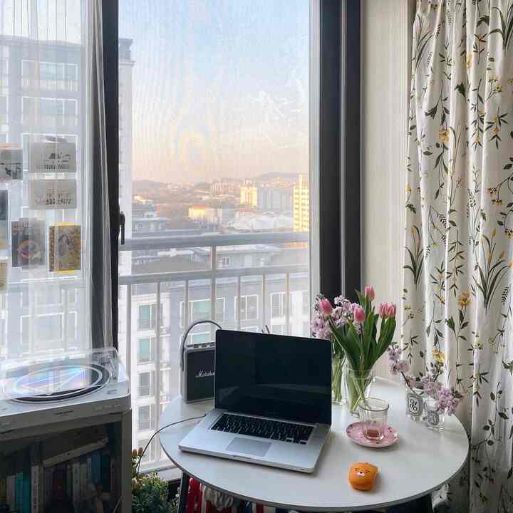 Bright white and pink toned small room featuring a round table with laptop and flowers, creating a cozy and luminous remote work space