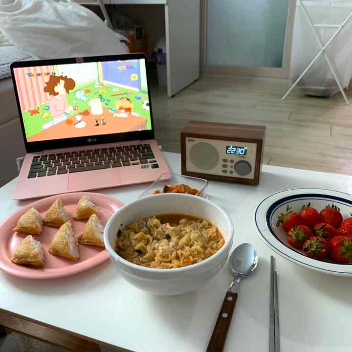 White-toned studio apartment dining space featuring laptop, ramen bowl, snacks, and Bluetooth speaker in cozy atmosphere
