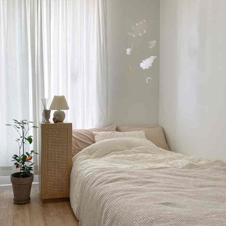 Bright natural-tone studio bedroom featuring beige bedding, cane storage cabinet, mobile decoration, and a potted plant creating a cozy atmosphere