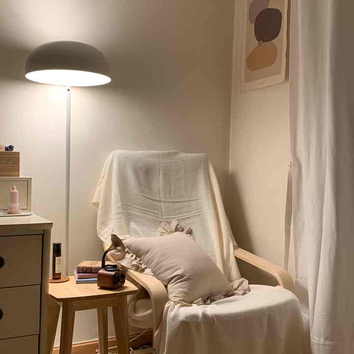 A cozy living room corner in white and wood tones featuring a comfortable armchair, stool, and warm floor lamp lighting