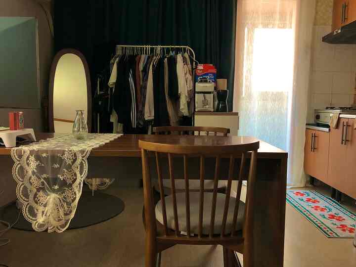 Natural-toned studio apartment dining space with warm wood furniture, lace curtains, and a bright balcony bringing in soft natural light