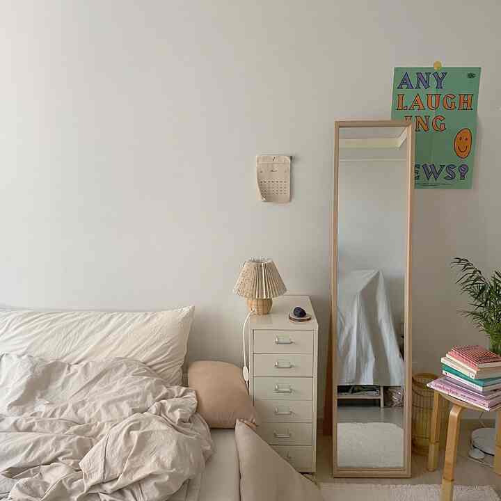 柔らかいナチュラルカラーの狭い一人寝室、ベッド横にチェストと全身鏡、木製スツールに本が積まれている印象的な空間