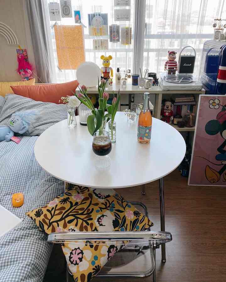 Bright toned small room for a single household featuring a white round dining table, transparent chair, curtains, plants, and a cozy home cafe vibe with brown wood flooring