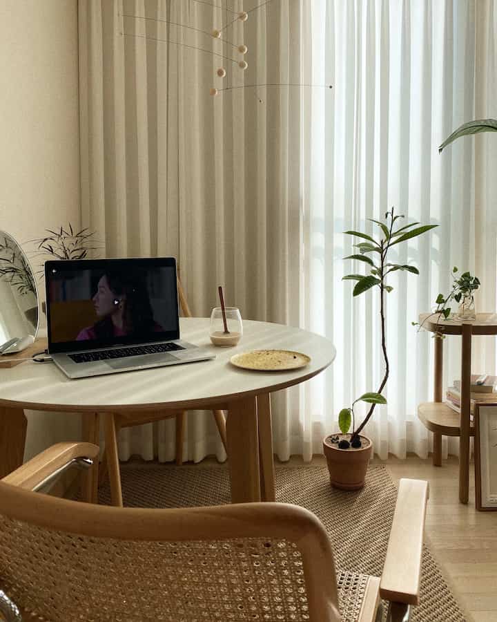 ナチュラルな木目調と白いカーテンが調和したホームオフィス兼ダイニングスペース、木製テーブルと椅子、植物が配置された落ち着いたインテリア