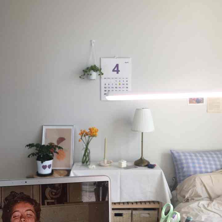 A white and natural color-toned bedroom combined with a home office, featuring a desk, lamps, and a calendar creating a cozy atmosphere.
