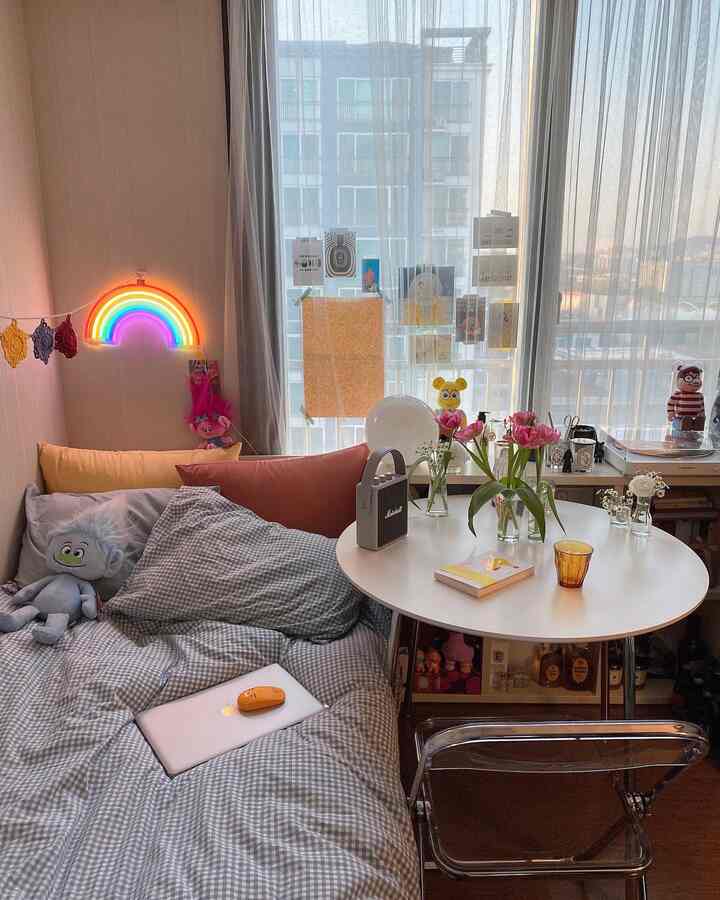 Soft beige and brown tone studio bedroom featuring checkered bedding, bright round table, vases with flowers, and a neon rainbow light creating a cozy atmosphere