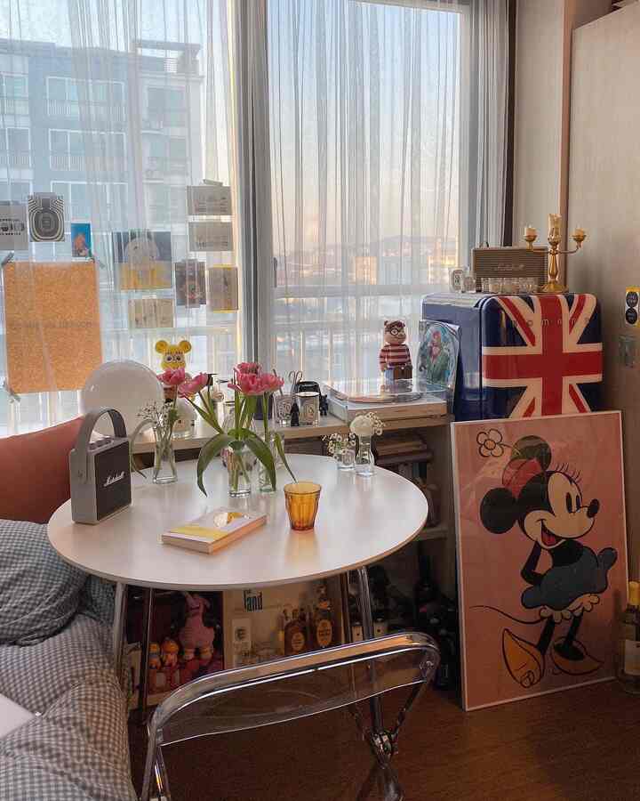 ウッドトーンのワンルーム、白い丸テーブルと透明な椅子を配置し、ミニ冷蔵庫とディズニーポスターが印象的な空間