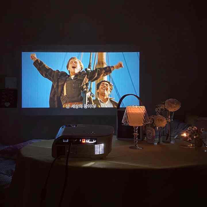 暗い夜のリビングで、黄色のテーブルクロスの上にプロジェクターとランプが置かれ、壁に映画が映し出された雰囲気のあるホームシネマ空間