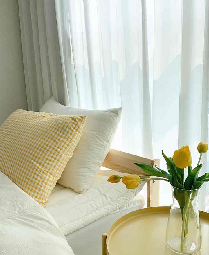 Bright natural white and yellow toned bedroom featuring wooden bed, checkered pillow cover, and cozy yellow tulip vase on nightstand